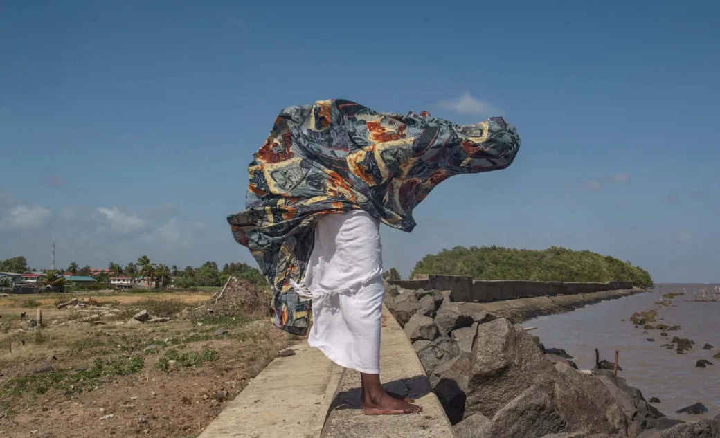 Black person in flowing clothing blowing in the wind holds arms out, face obscured.