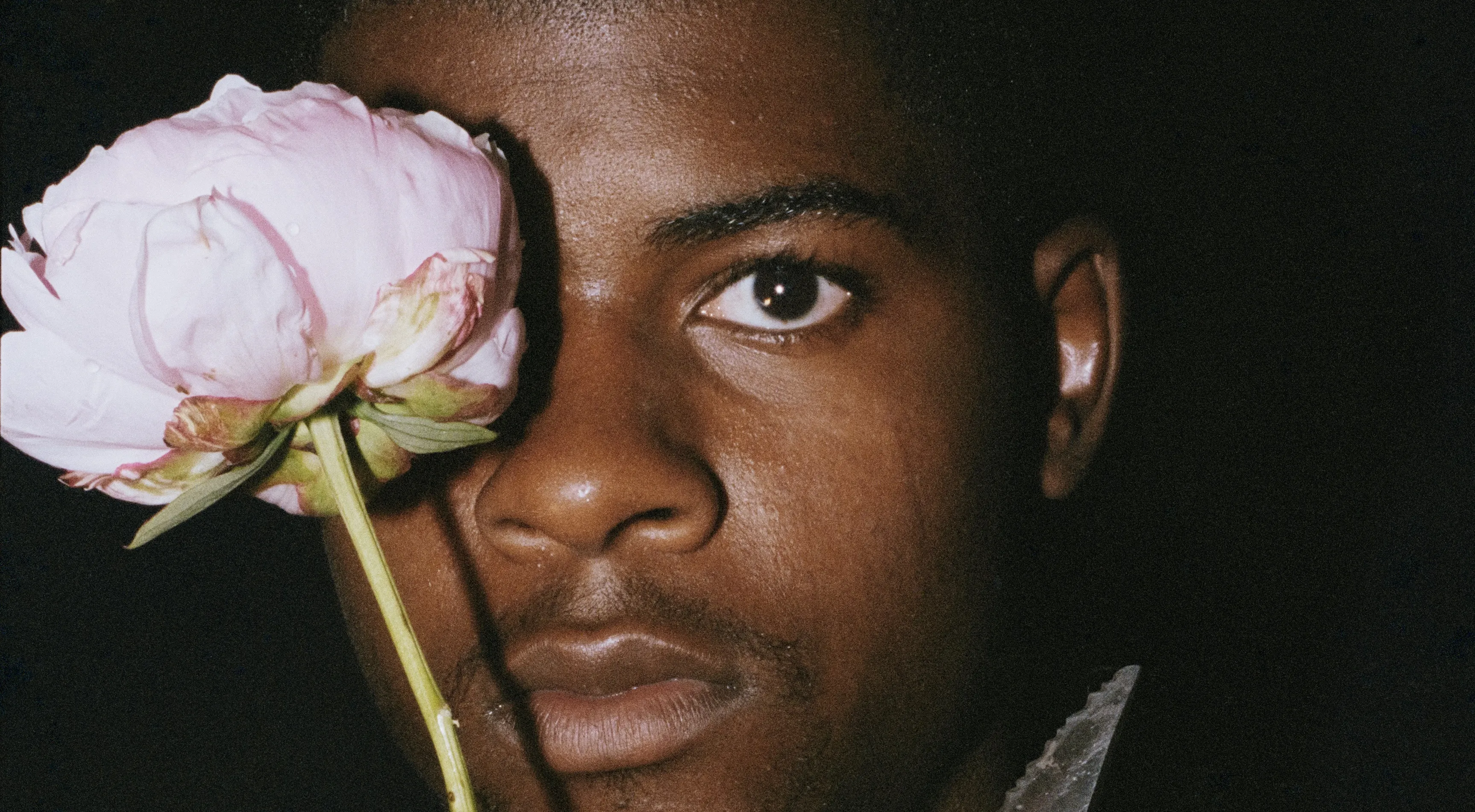 A man in the dark, with a flower raised and covering one eye. 