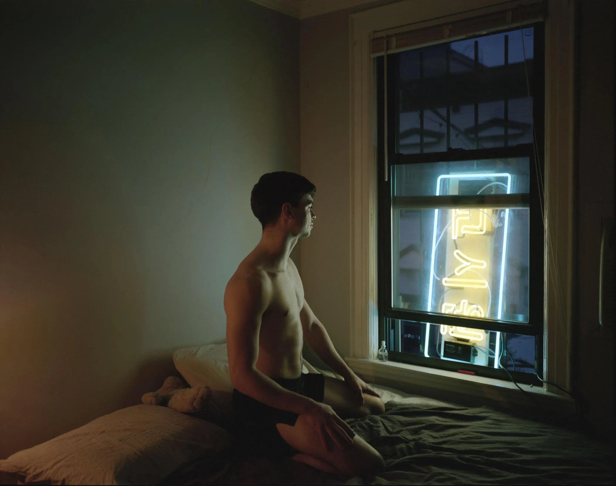 A man sitting on his bed, looking at a blue and yellow neon sign glowing outside of his bedroom window.