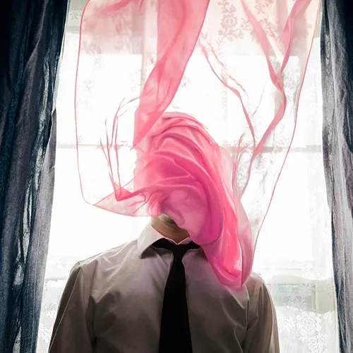 A man standing in front of the bright window, an almost transparent pink curtain wrapped around his face.